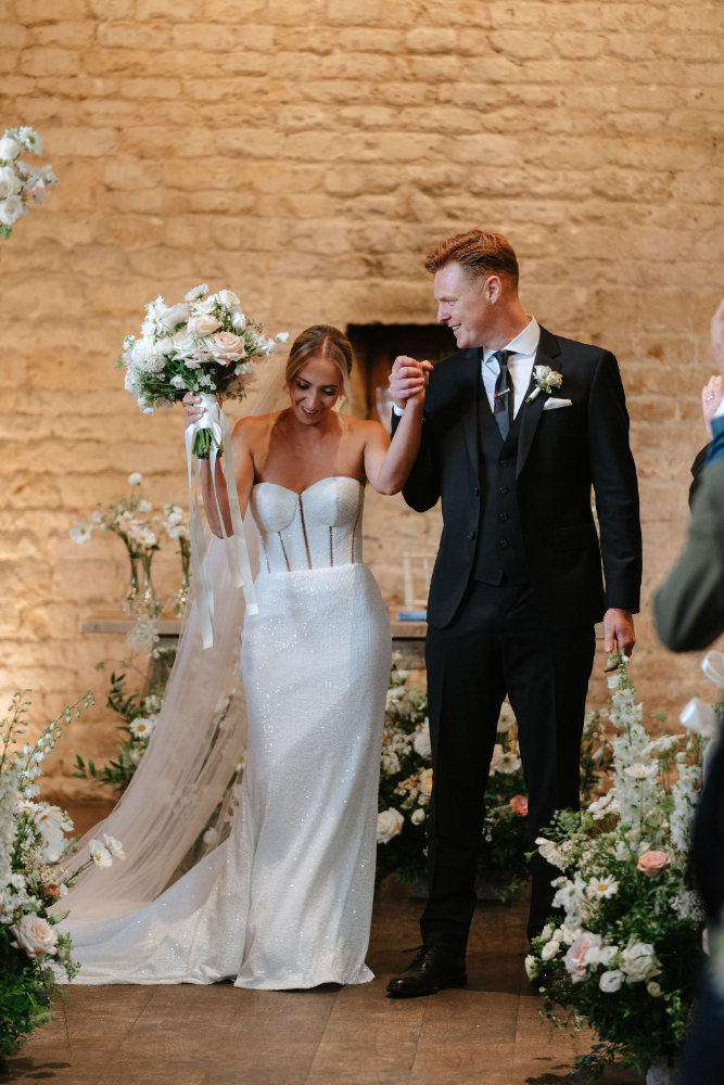 A bride and groom celebrating walking back down the aisle after their ceremony at Lapstone Barn, the aisle is filled with whimsical flowers by Sally Gill Flowers.